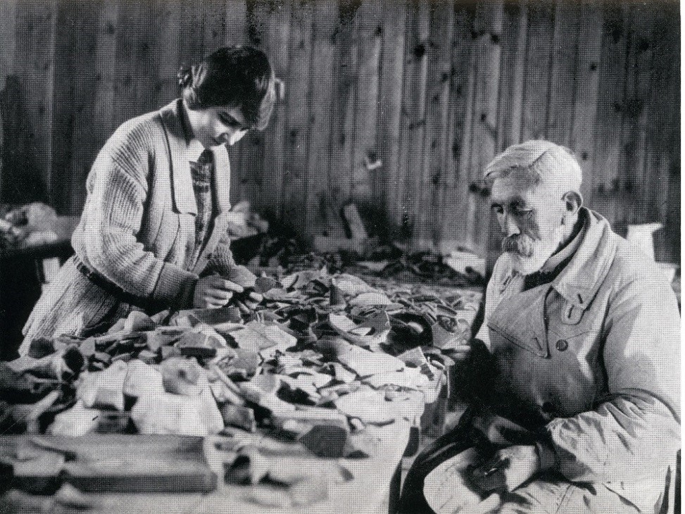 alt="woman and man looking at piles of pottery on either side of a long table"