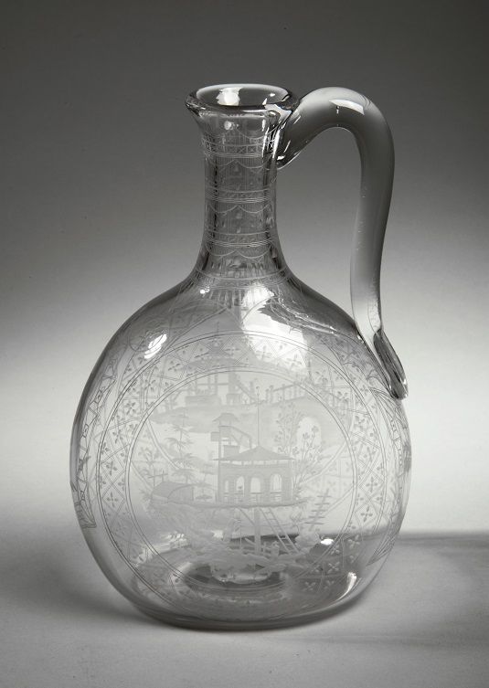This single-lipped decanter is made from clear glass with an applied handle. It has finely engraved buildings in a landscape