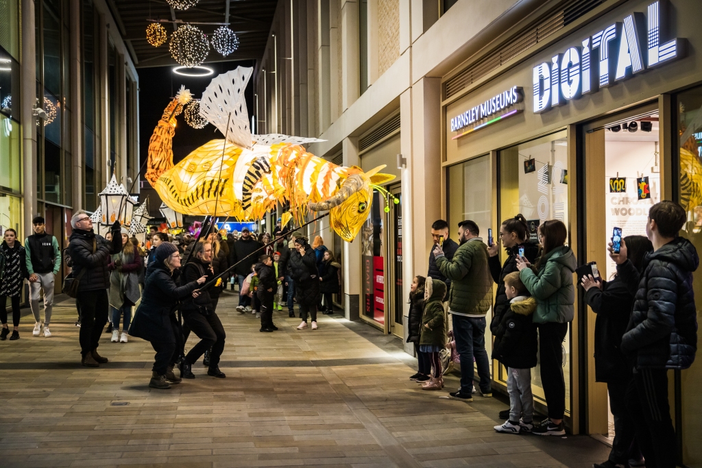 Eldon Beast puppet interacting with a small crowd in front of the Barnsley Museums digital space in the Glassworks shopping centre. People are taking photographs. The beast is a large illuminated puppet with a rabbit's head, a tiger's tail, slug horns and wings. It is held above head height on poles and operated by four people standing underneath. It looks like it is bowing it's head to look at two small children standing with two adults. The children are looking up in wonder.