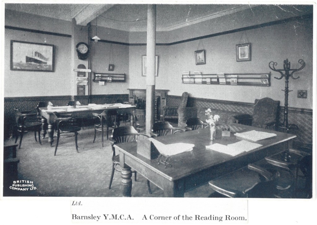 Photograph entitled ' Barnsley YMCA a corner of the reading room' It shows a room with tables and chairs, with a clock and racks for newspapers on the wall. Other features include a painting of a steam ship and a hat stand, and a corner fireplace. The decoration looks very smart, with a wooden dado rail running along the walls. 