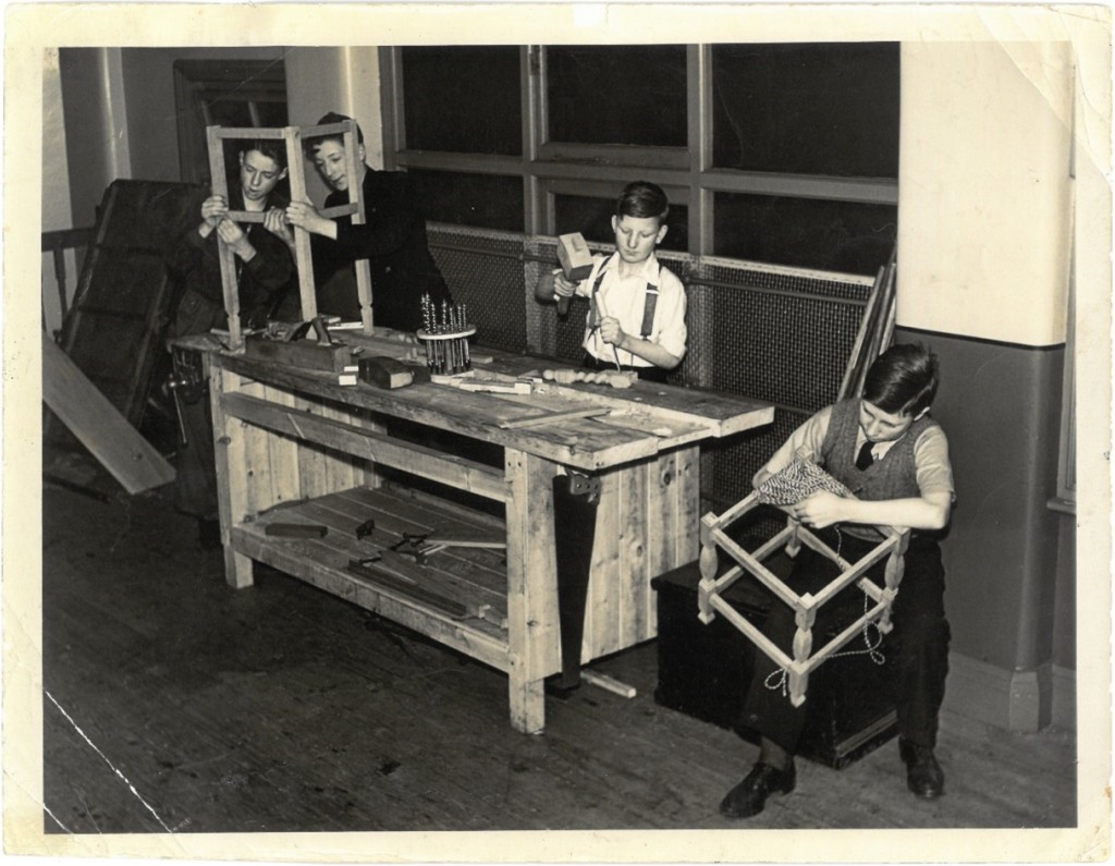 Photograph of four boys in a wood working class. They are all working at a wooden work bench which has a range of tools on it, including a plane and chisels. Two boys on the left are fitting the legs and cross supports together, a boy in the middle is working on a turned spindle with a hammer and chisel, and a boy on the right is polishing the legs of a chair (or stool). 