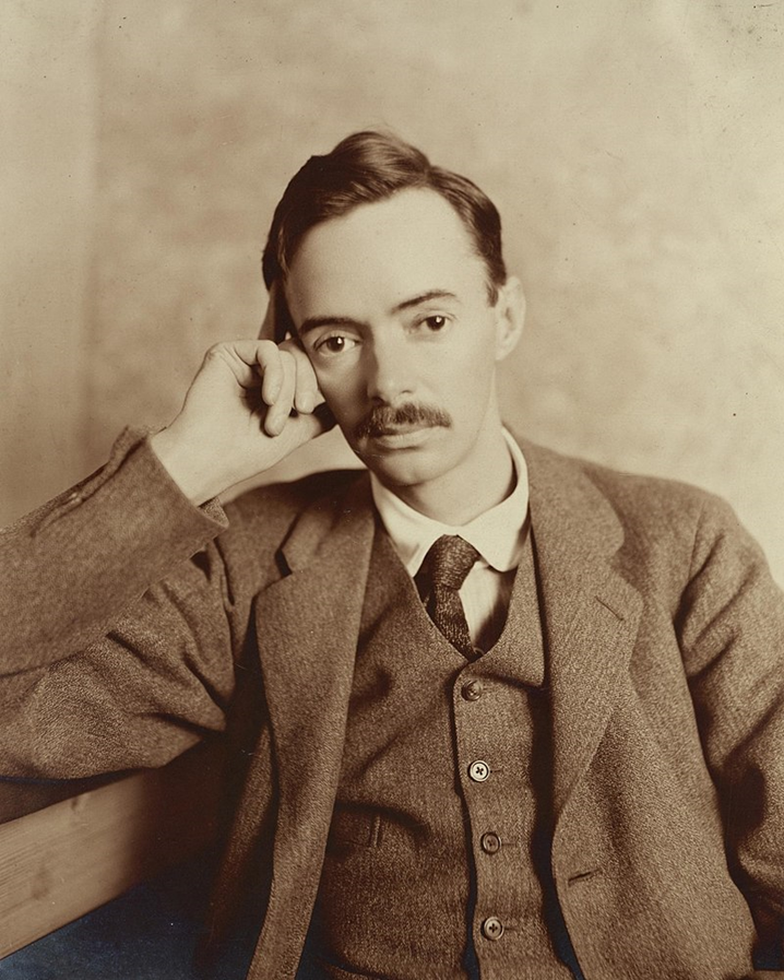 A picture of Ernest wearing a suit and tie. His right hand is resting against his head. He has short hair and a moustache.