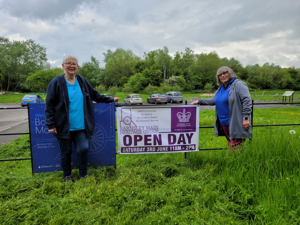 Val and Sarah stood on the outskirts on the site on  grass and between them is a large sign for an open day event. Behind them is the car park.